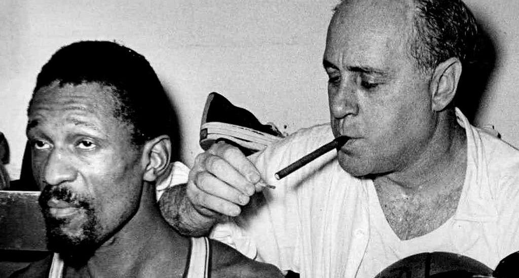 Auerbach smoking a cigar behind Bill Russel after winning the 1966 NBA Finals. (Via Wikimedia Commons)